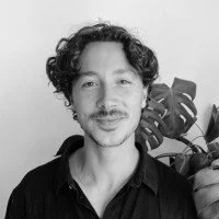 A man with curly hair and a mustache smiling at the camera, wearing a dark shirt, with large leaves in the background.