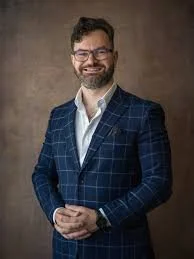 A man with glasses, a beard, and short hair, dressed in a checkered blue blazer over a white shirt, smiling against a neutral background.