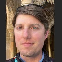 A young man with light skin and brown hair, wearing a patterned shirt, standing outdoors with decorative stone architecture in the background.