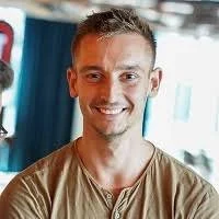 A woman with short blonde hair, blue eyes, wearing a dark blazer and light blouse, smiling and standing indoors.