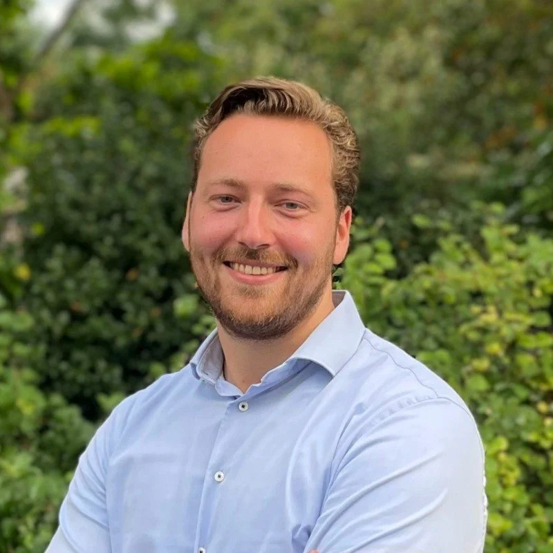 A smiling man with light brown hair and a beard, wearing a light blue button-up shirt, standing outdoors with green trees in the background.