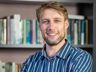 A man with light brown hair and a beard smiling, wearing a blue and white striped shirt, standing in front of a bookshelf filled with books.