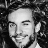 A black and white close-up portrait of a smiling man with dark hair and a beard, facing forward.
