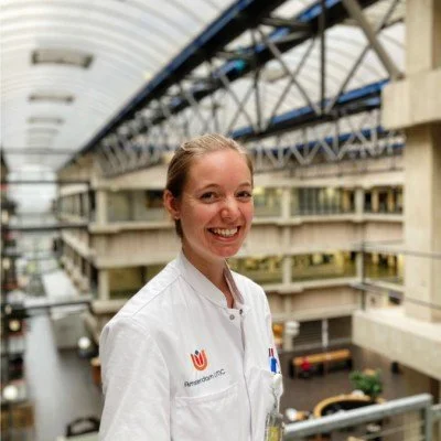 Portrait of a middle-aged woman with short blond hair, wearing a navy blazer and earrings, standing and smiling in an indoor setting.