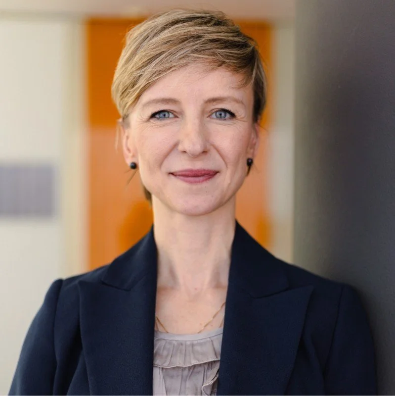 Woman with long dark hair wearing a blue sleeveless top and large blue earrings, sitting on a dark-colored couch against a plain background.