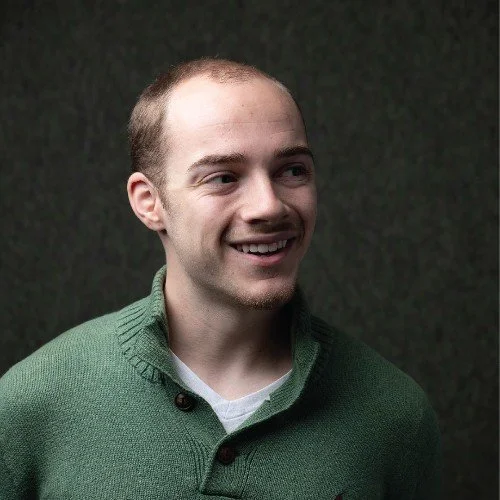 A young man with short hair and a slight smile, wearing a green sweater, standing in front of a dark background.