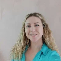 Man with a beard and short hair, wearing a gray long sleeve shirt, sitting and smiling outdoors with green bushes in the background.