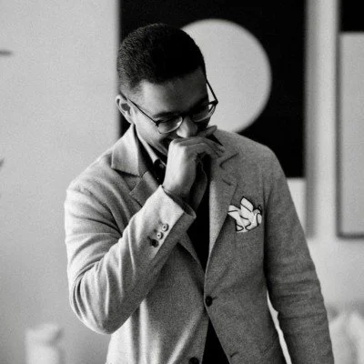 A young man with glasses, wearing a blazer with a pocket square, smiling and covering his mouth with his hand in an indoor setting.