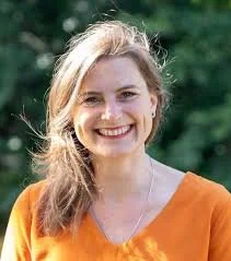 A woman with long blonde hair smiling outdoors, wearing an orange top.