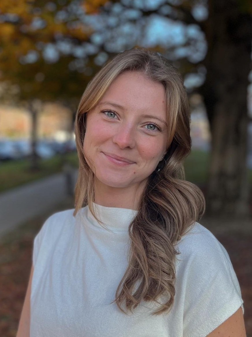 A young woman with blonde hair and blue eyes smiling outdoors, wearing a white top, with trees and a park in the background during daytime.
