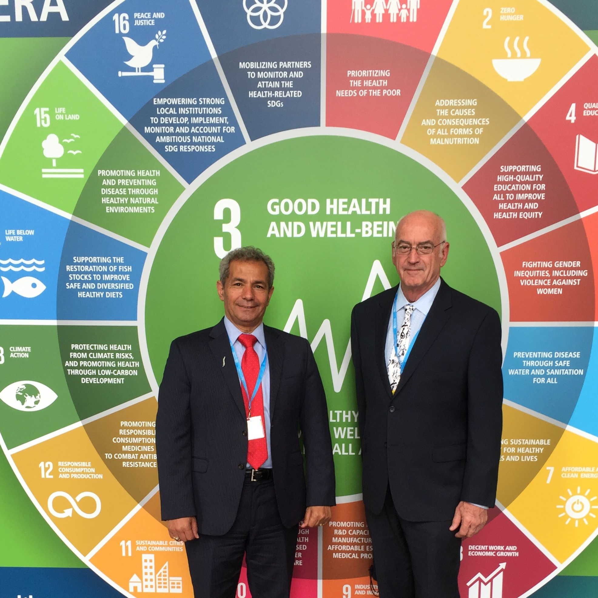Two men in business suits standing in front of a colorful circular infographic displaying the United Nations Sustainable Development Goals, with the central goal 'Good Health and Well-Being' highlighted.