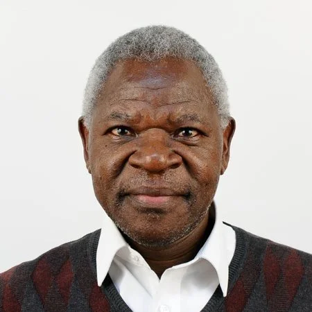 Close-up portrait of an elderly man with gray hair, wearing a white shirt and dark sweater, against a plain white background.