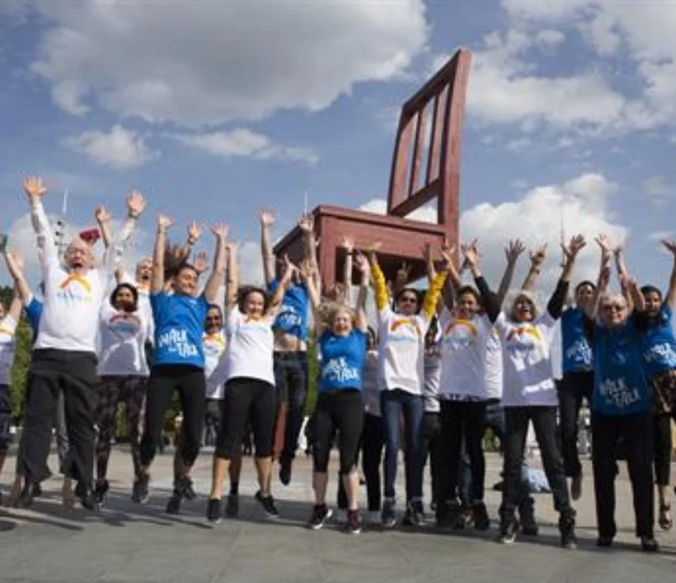 Group of people standing outdoors under a partly cloudy sky, raising their arms, with a large wooden chair sculpture in the background.