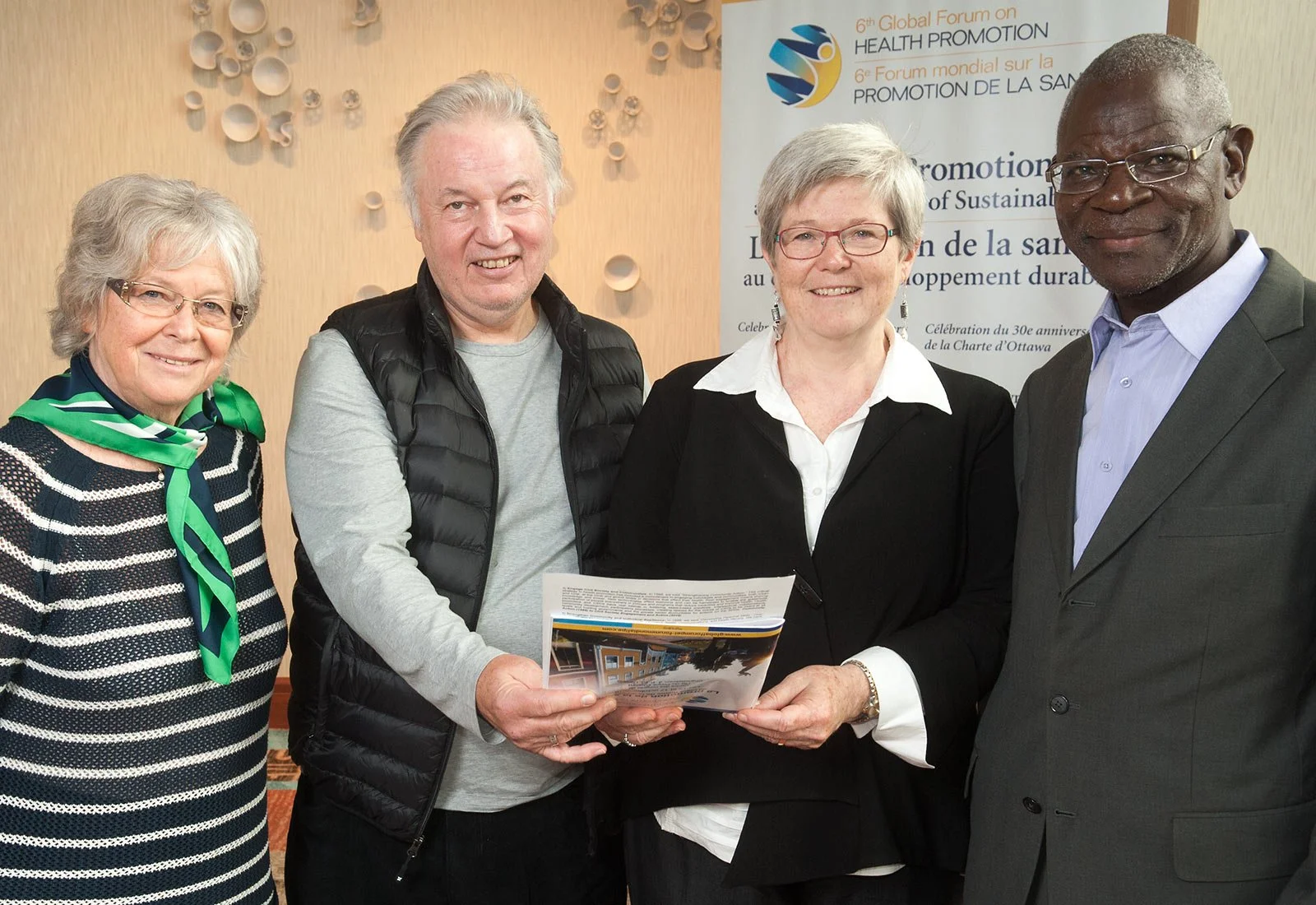 Four people standing together at a conference, holding a brochure, with a banner in the background that reads '6th Global Forum on Health Promotion'.