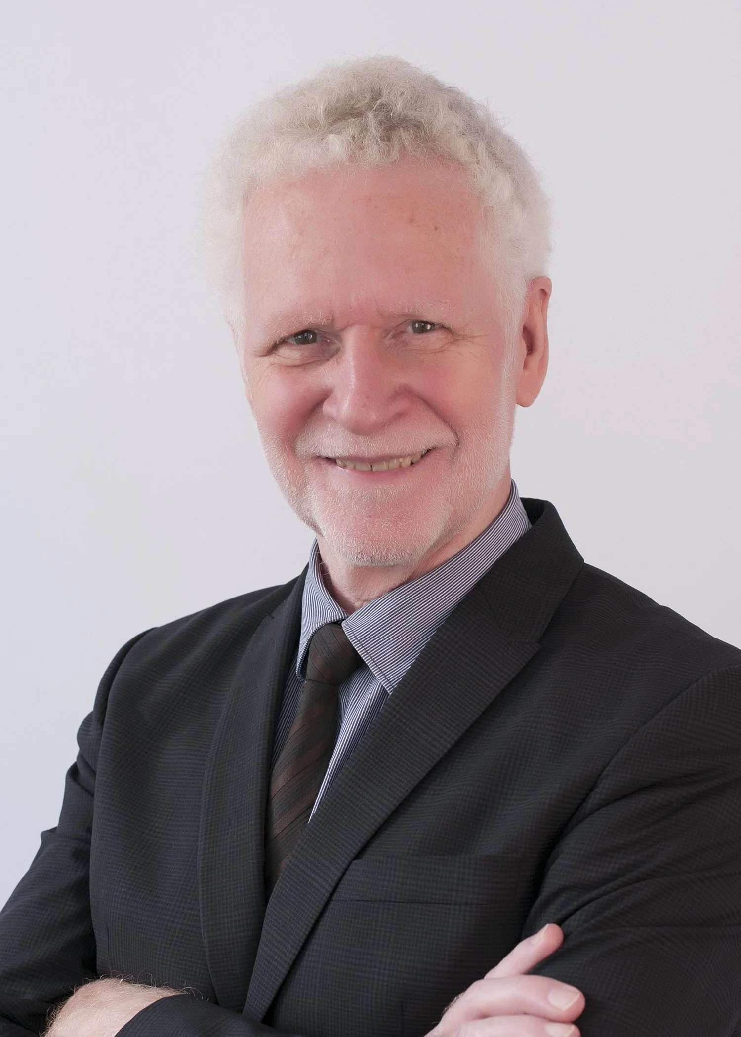 A smiling older man with white hair and a beard in a black suit, striped shirt, and dark tie, standing against a plain light background.
