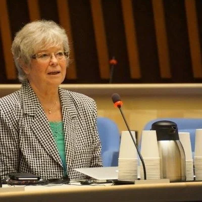 An elderly woman with short gray hair, glasses, and a checkered blazer sitting at a table with a microphone, cups, and papers, speaking at a formal event or meeting.