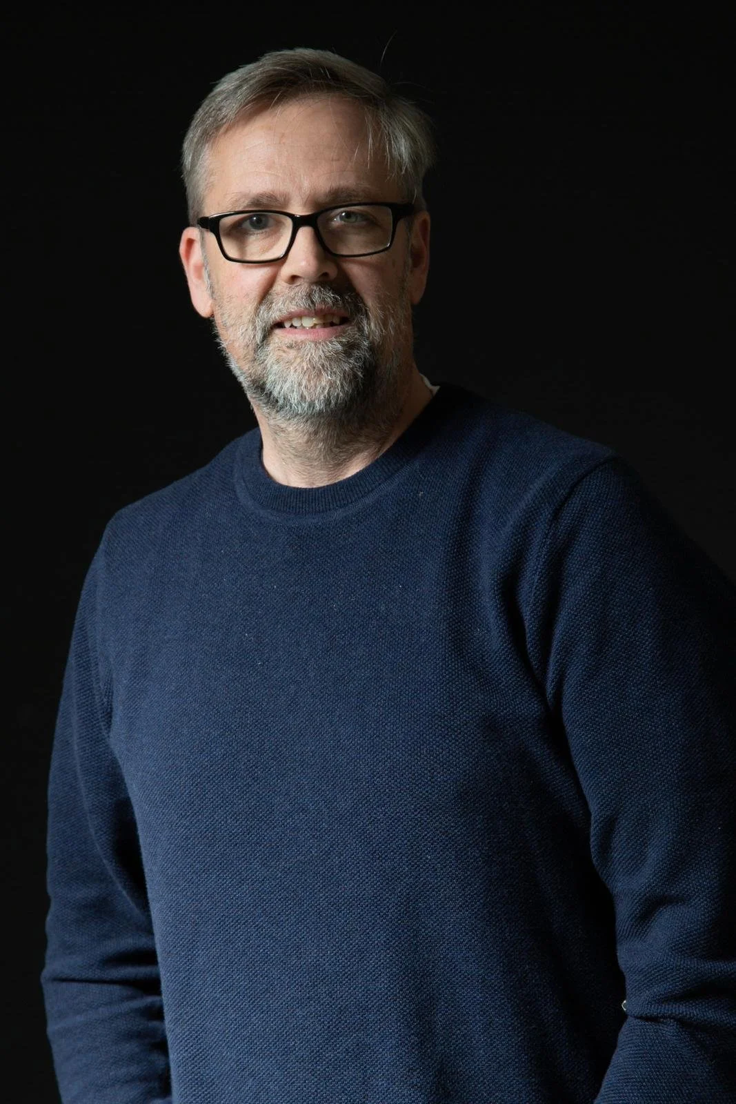 A middle-aged man with glasses, gray hair, and a beard, wearing a navy blue sweater, posing against a black background.