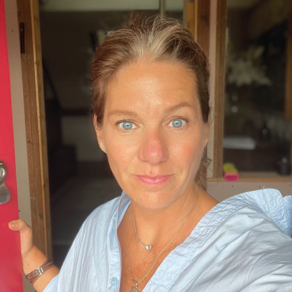 A woman with blonde hair and blue eyes standing at a doorway, wearing a light blue top, with a background of a wooden interior.