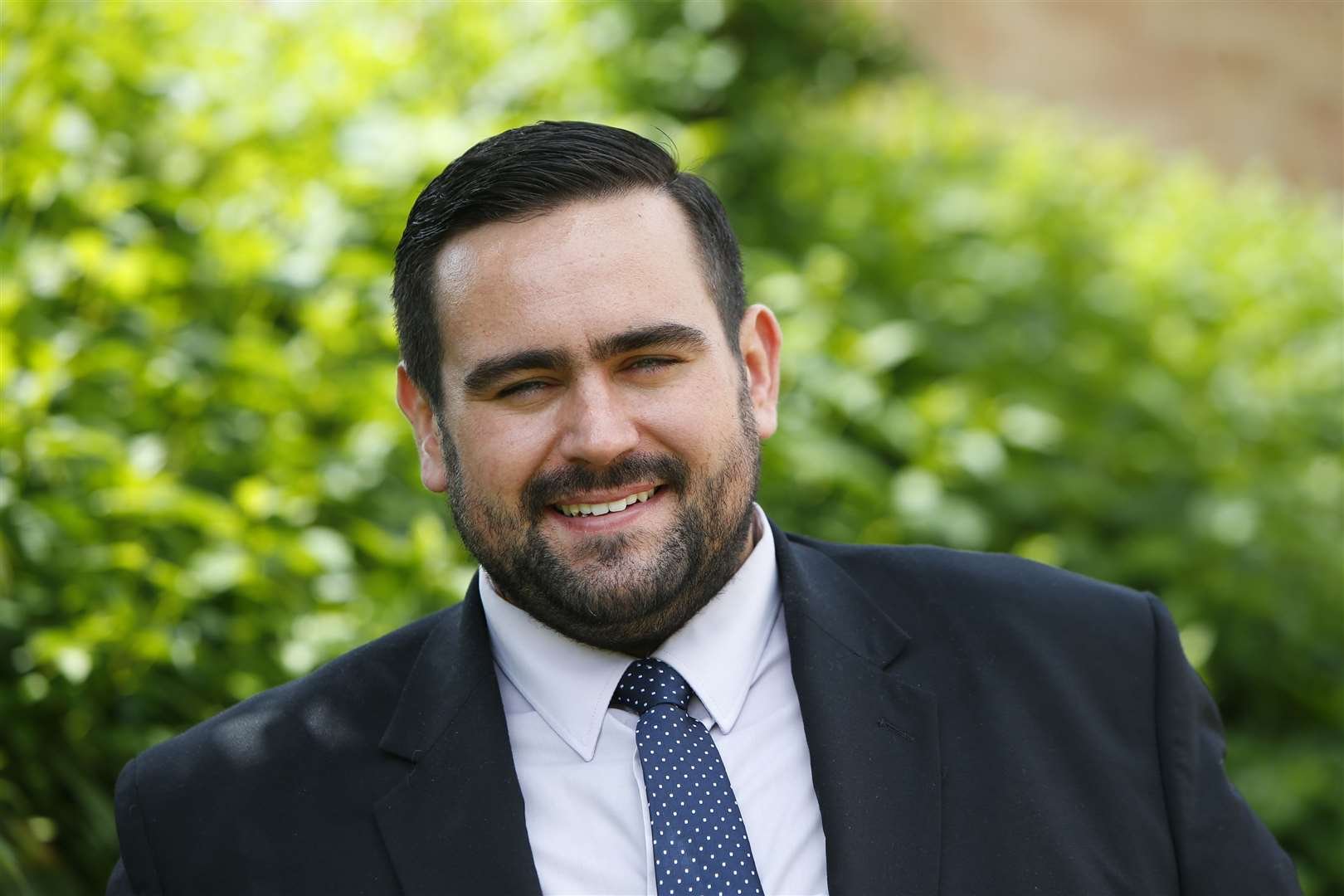 A smiling man with dark hair and a beard in a black suit, white shirt, and blue polka-dot tie, standing outdoors with green foliage in the background.