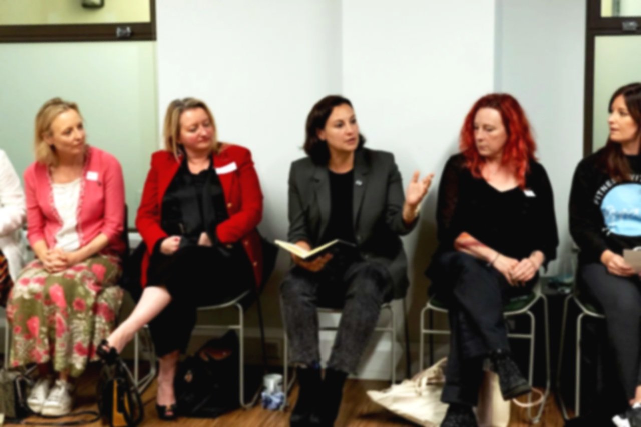 Group of five women sitting in a row on chairs in a meeting room, engaged in conversation.