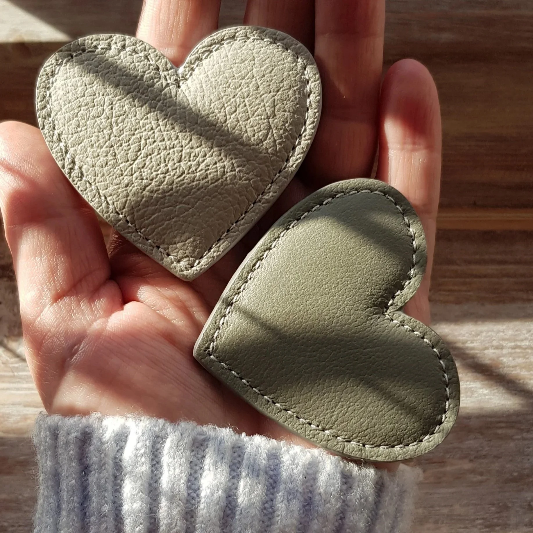 A hand holding two small leather heart-shaped objects, one beige and textured, and the other smooth and olive green. Sunlight casting shadows on the objects.