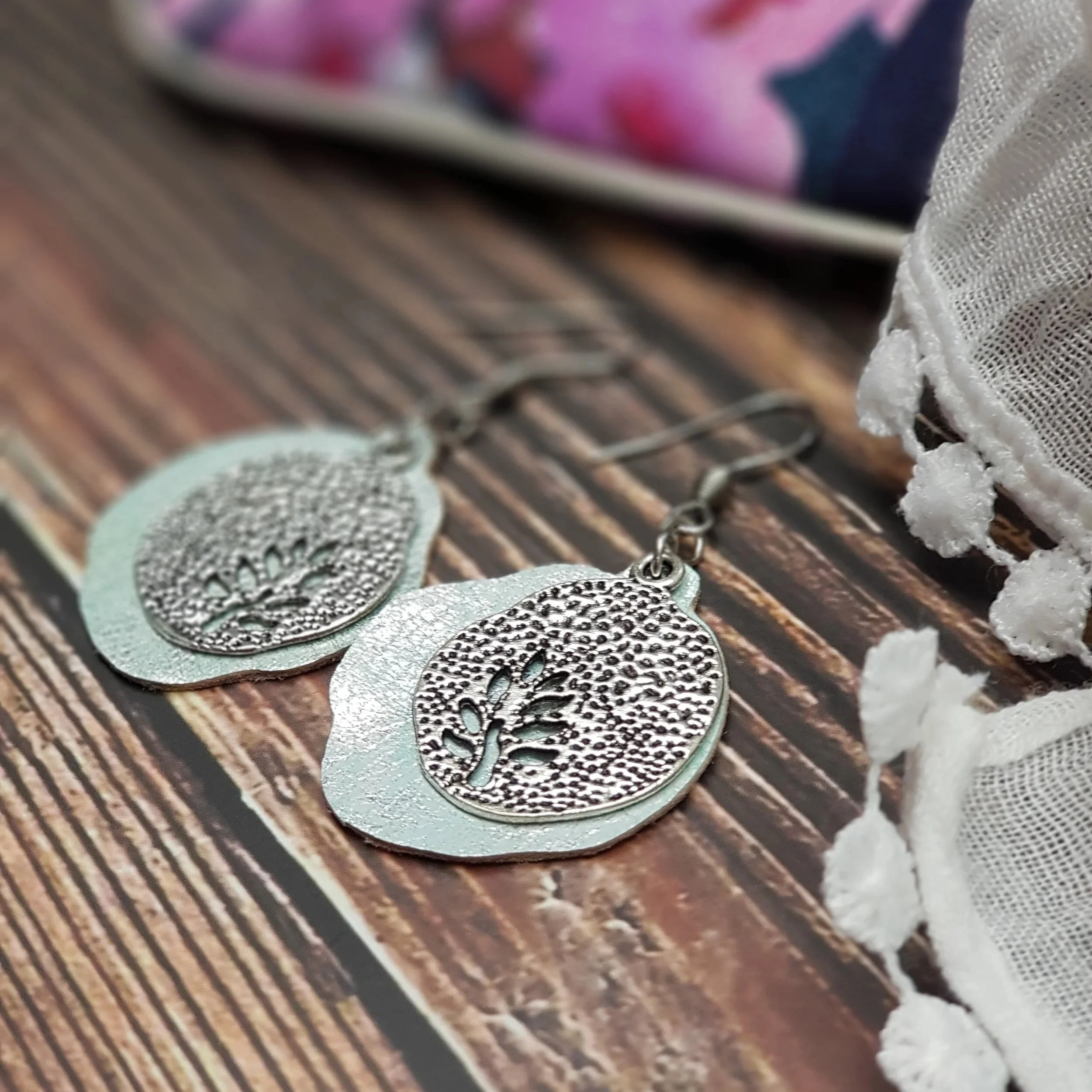 Pair of silver earrings with tree of life design resting on a wooden surface near a lace fabric.