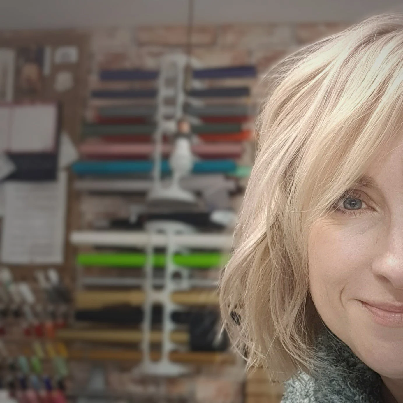 Close-up of a blonde woman's face, smiling slightly, with a blurred background of colorful objects and shelves.