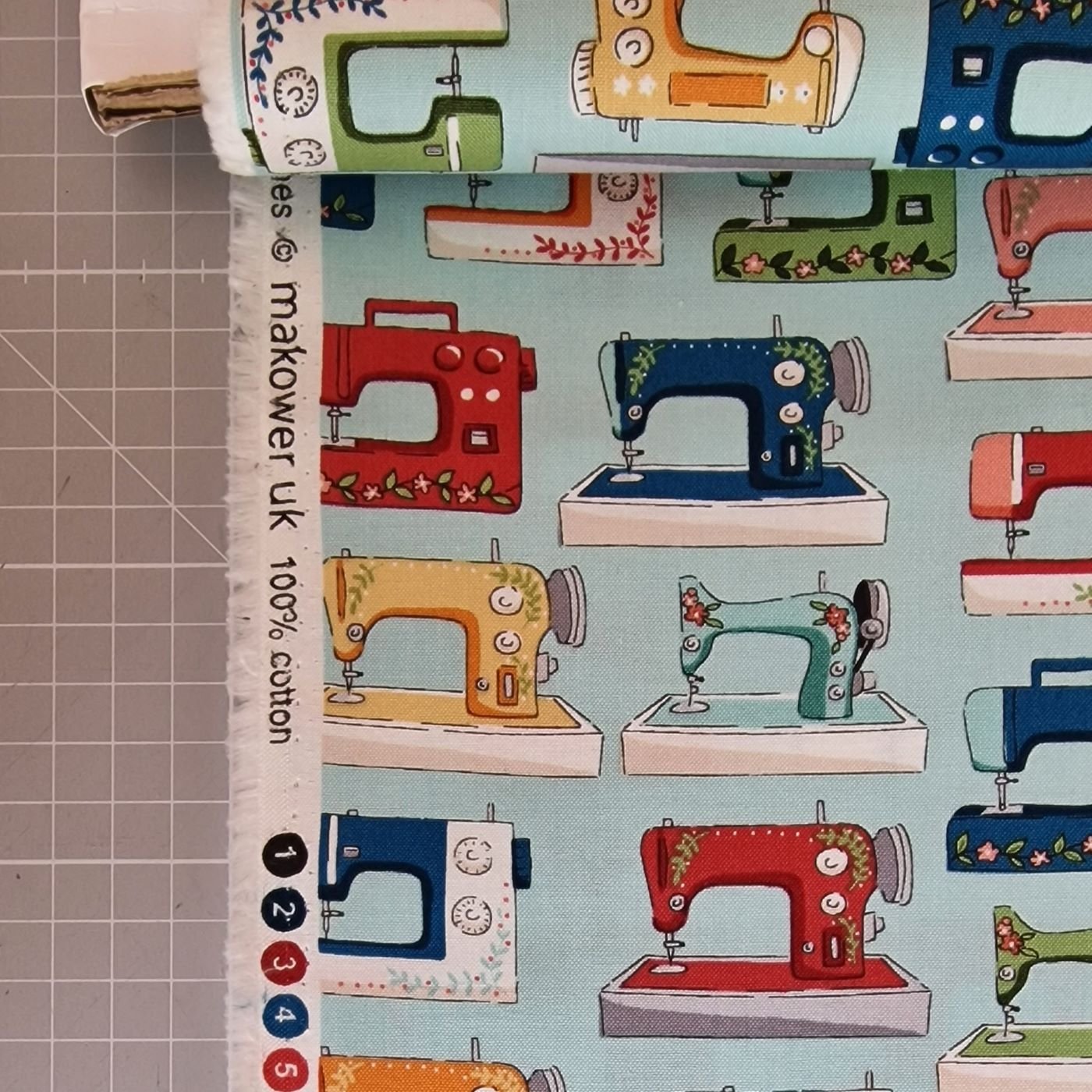Bolt of cotton fabric with blue background and pattern of colourful sewing machines. It's by Makower and Sewing Room collection