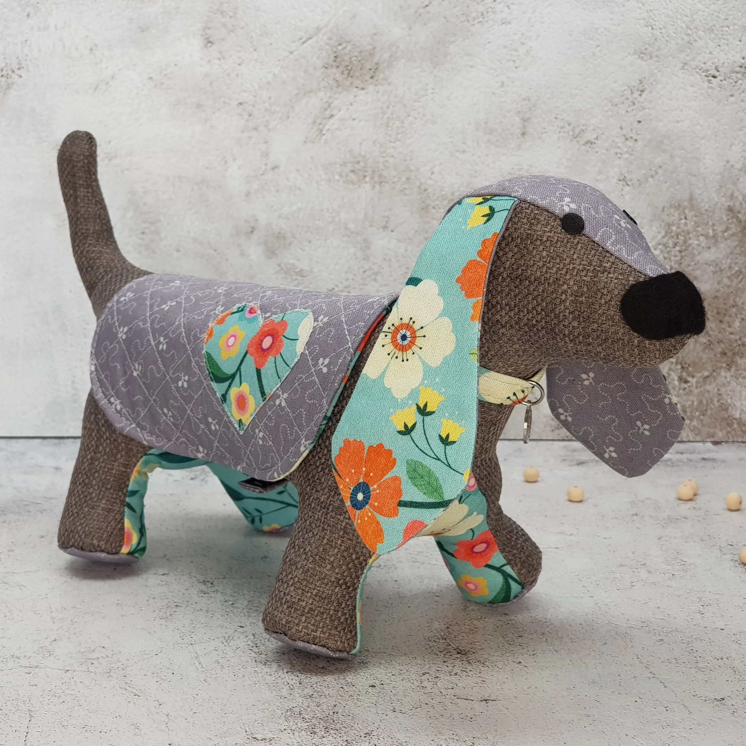 A fabric stuffed toy dog with a floral pattern and heart design on its back, standing on a light textured surface against a gray wall.