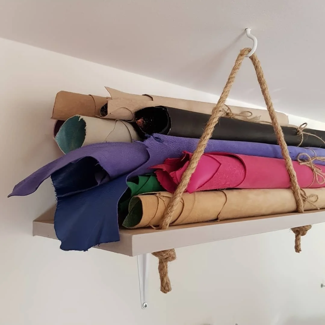 Shelf holding rolls of colorful fabric and paper, suspended by a rope hanger from the ceiling.