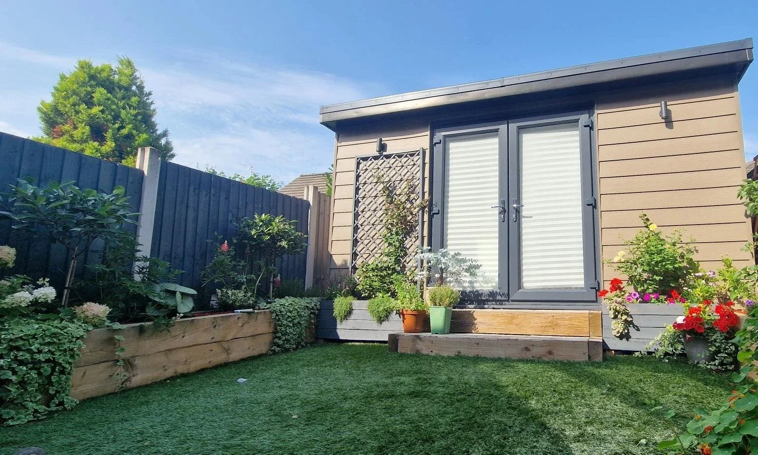 A backyard garden with a small shed, potted plants, and a wooden deck, surrounded by a blue fence and green grass.