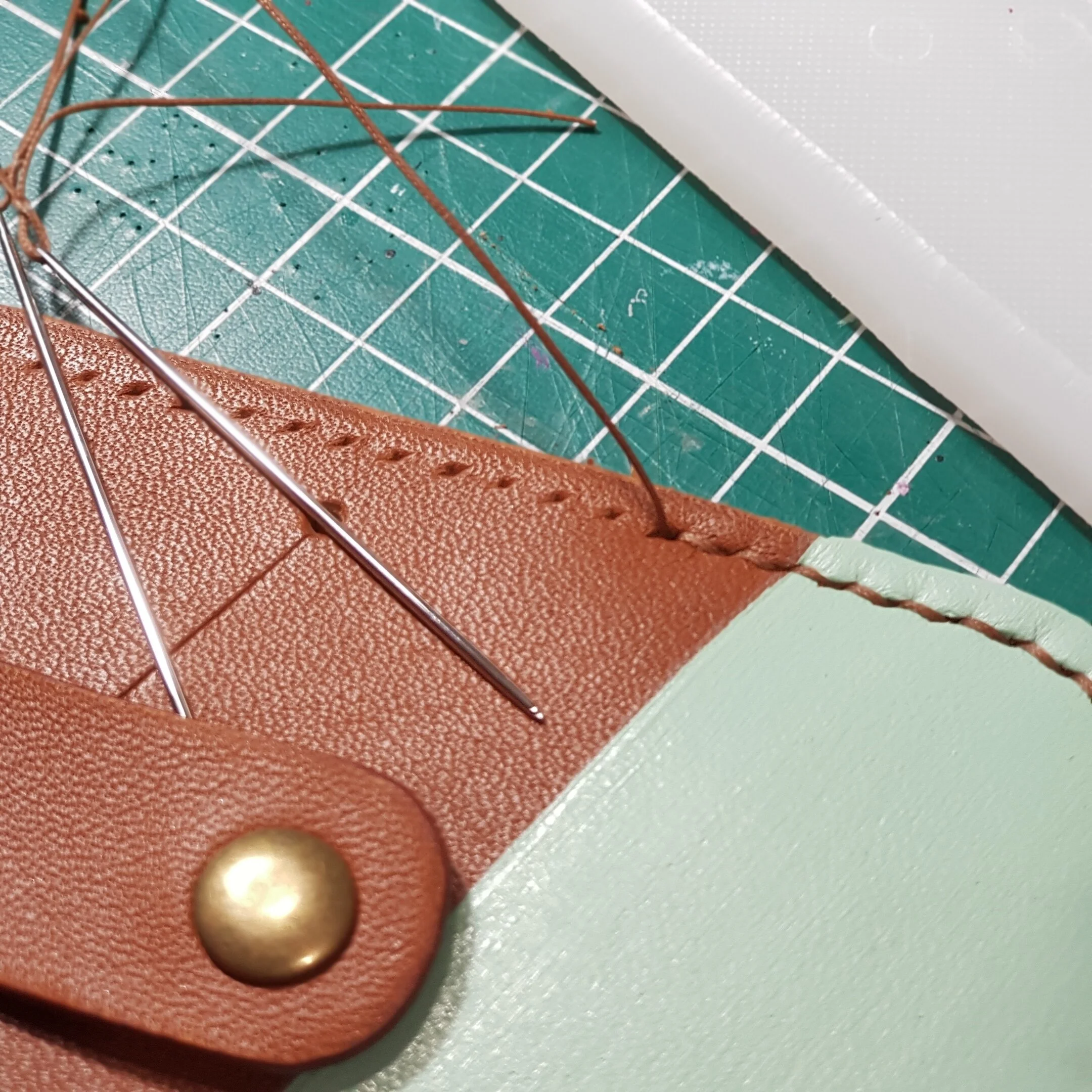 Close-up of a leather crafting project with a piece of brown leather, metallic pins, and stitching on a green cutting mat.