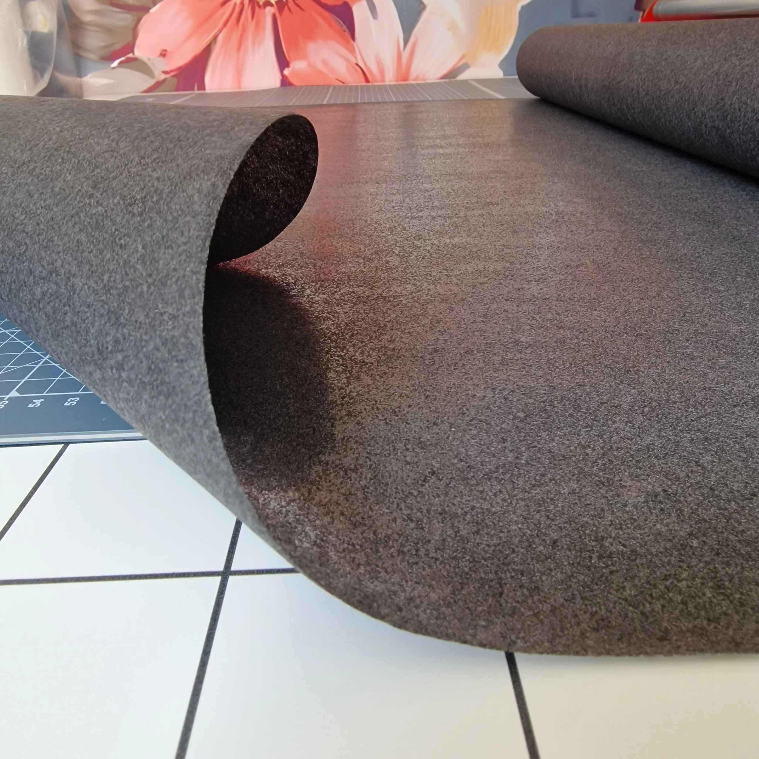Close-up of a roll of dark gray felt fabric partially unrolled on a cutting mat with measurement grid, with a colorful flower mural in the background.