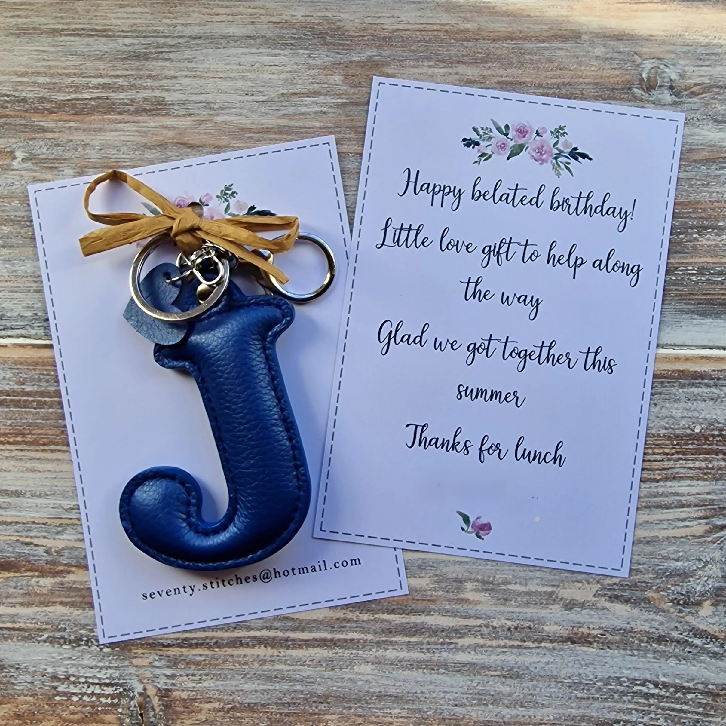 A blue letter 'J' leather keychain attached to a metal ring, next to a birthday note with floral decoration, on a wooden surface.