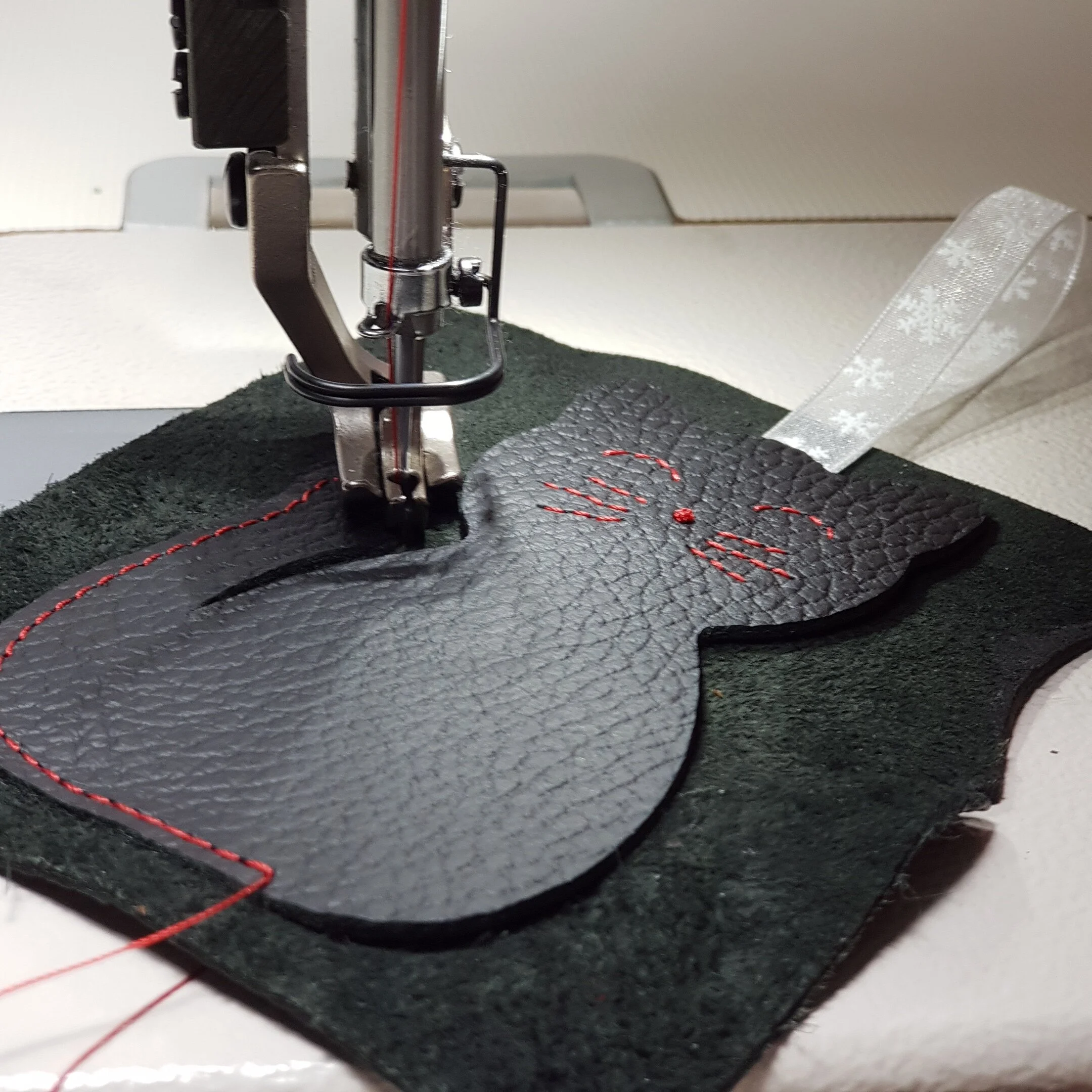Close-up of a leather mitten being embroidered with red thread, held in place on an embroidery machine with a dark fabric background.
