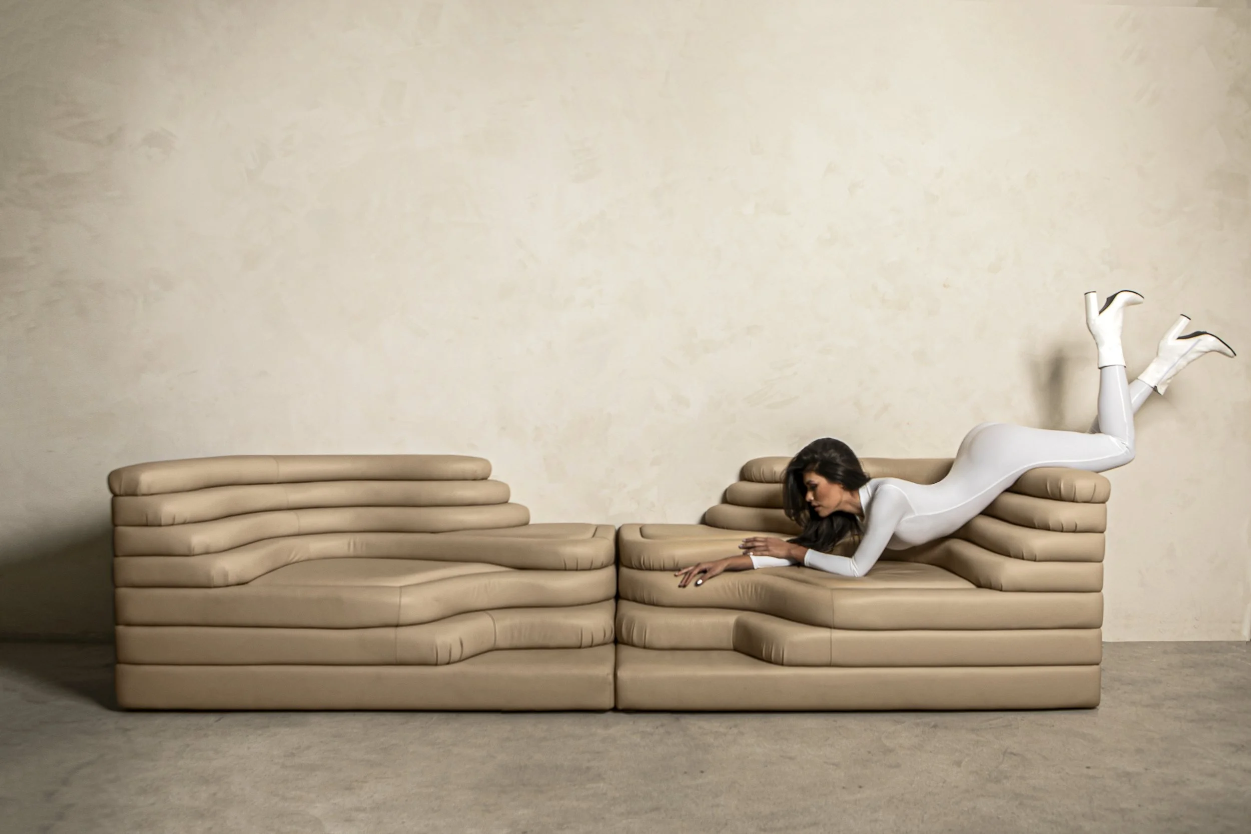Woman dressed in a white bodysuit lying on front on a beige sofa with her legs raised and crossed at the ankles, against a plain beige wall.