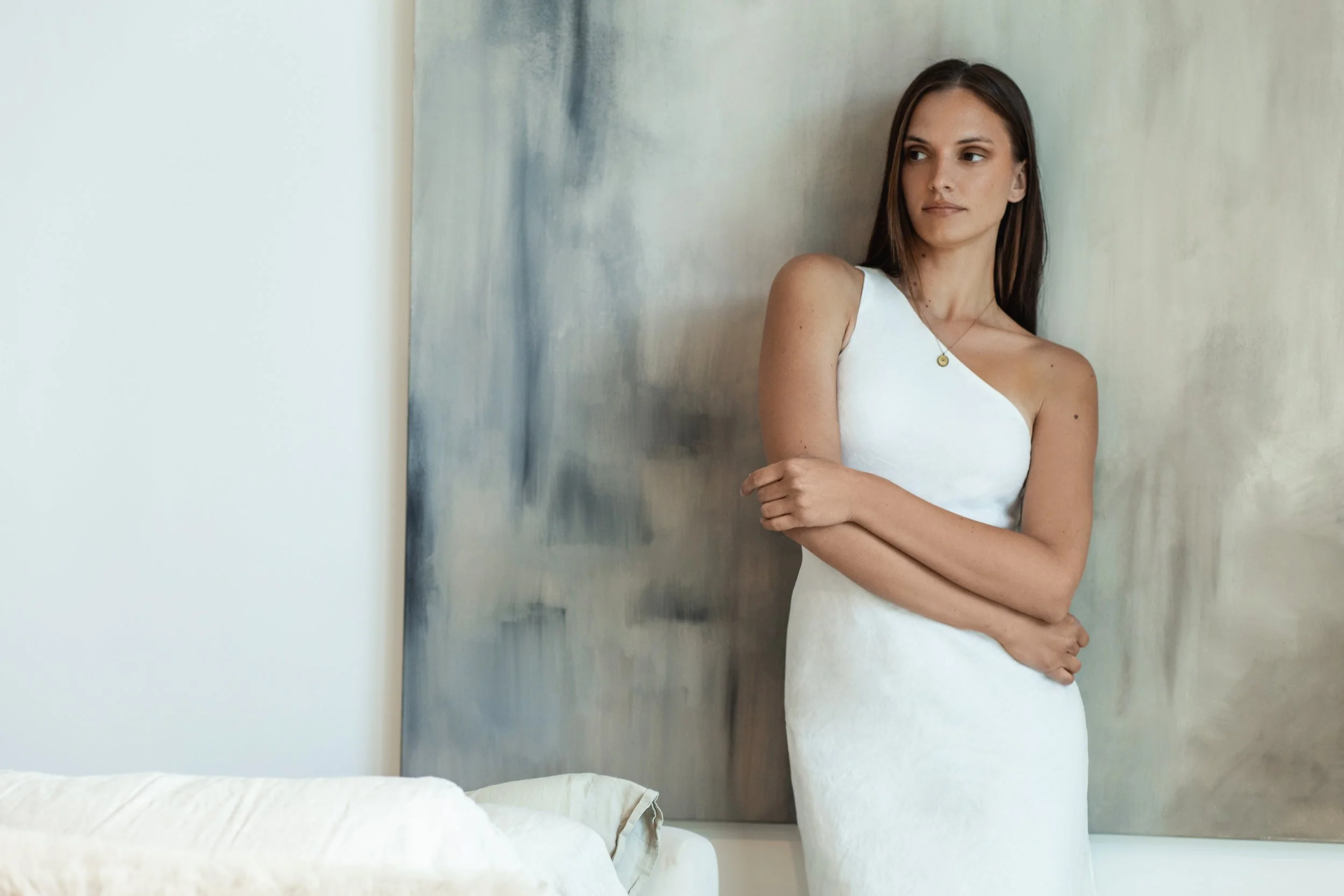 A woman with long dark hair wearing a white one-shoulder dress, standing with arms crossed against a textured wall with a painting in the background.