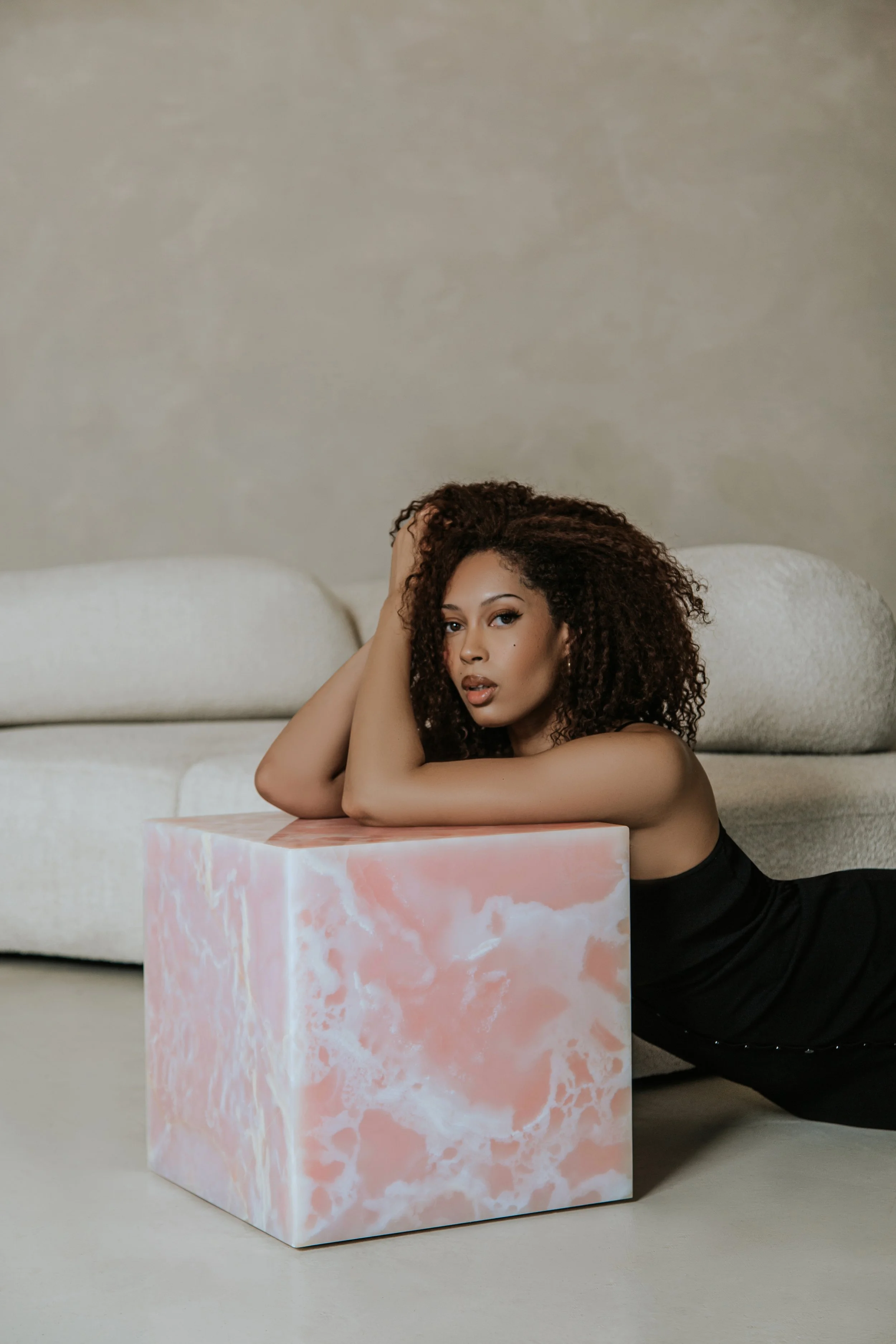 A woman with curly hair lies on the floor, resting her head on her hand, in front of a pink and white marble-patterned cube, with a beige sofa and plain wall in the background.
