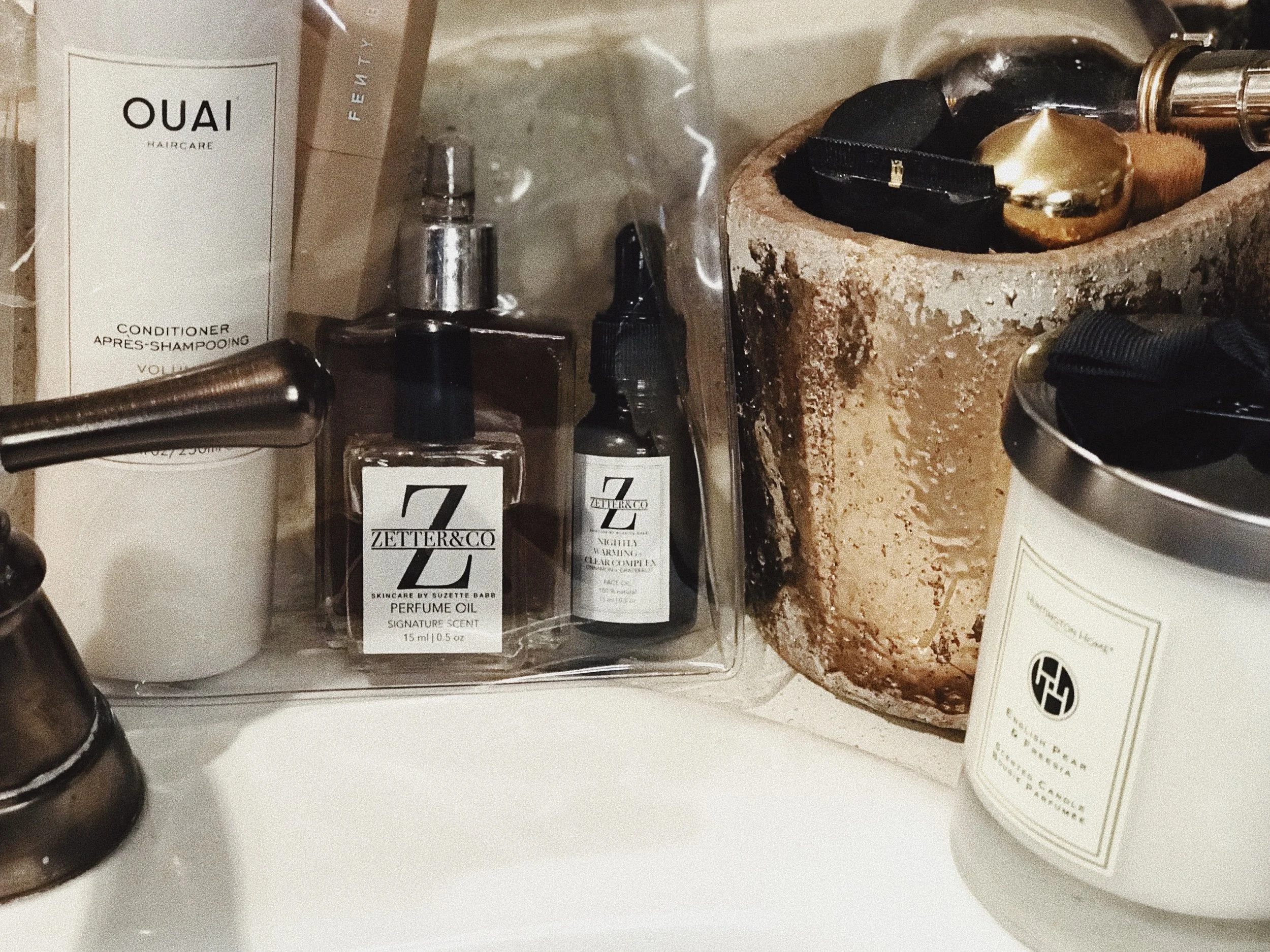 A collection of personal care and beauty products, including Ouai haircare conditioner, several perfume bottles, and a round container with a rough, textured surface.