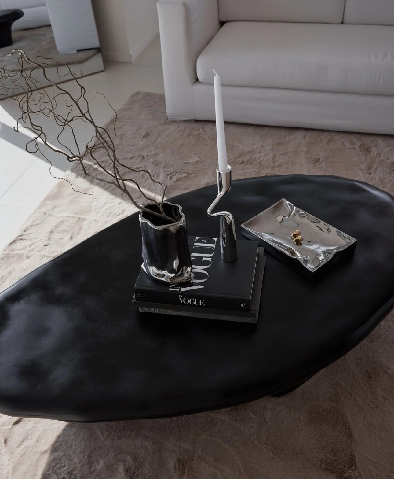 Black coffee table with a silver vase holding twisted branches, a silver candlestick with a white candle, two stacked magazines titled 'Vogue' and 'Women', a rectangular silver dish with a small gold ring, and a larger woven tray, in a living room wi