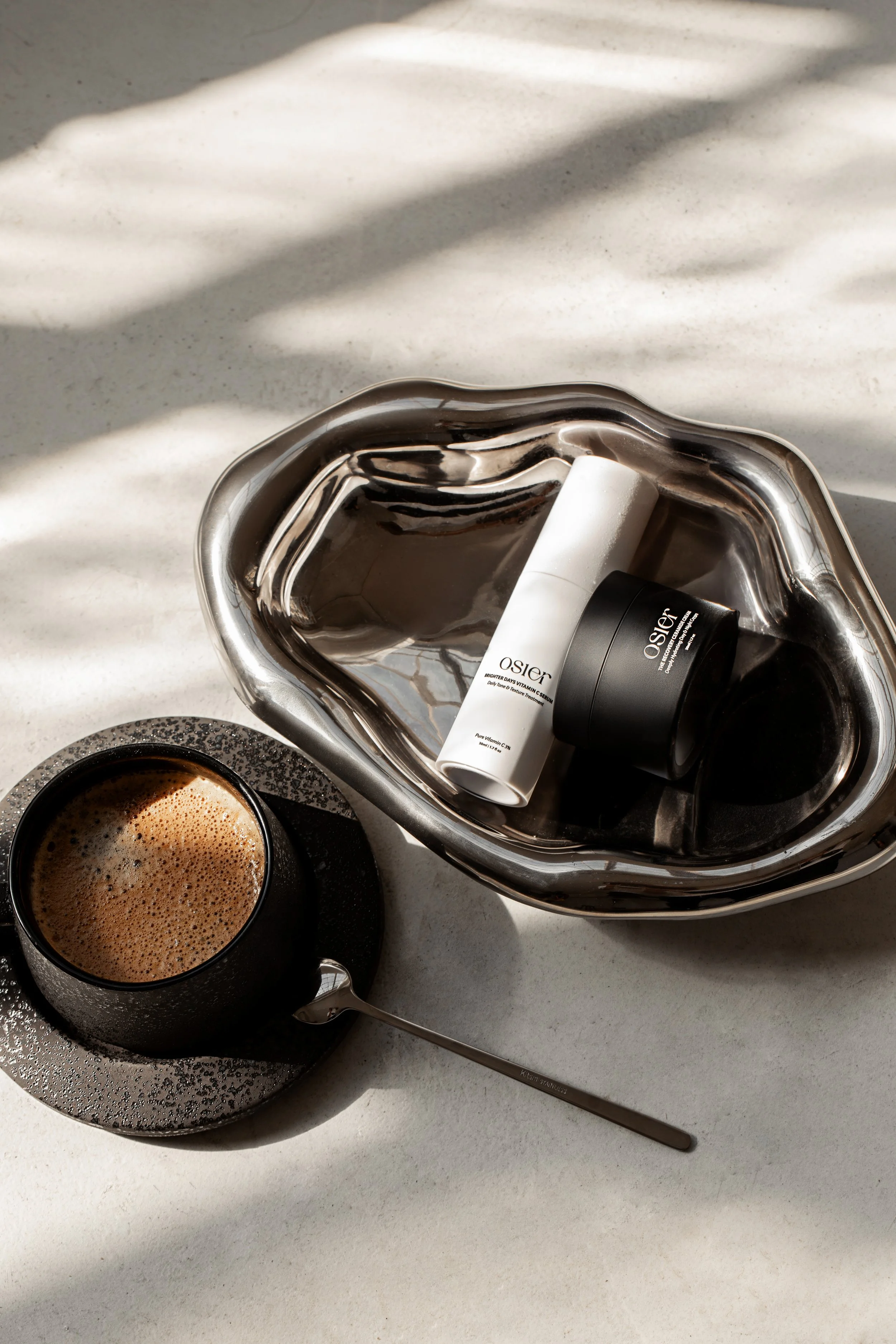 A cup of coffee on a saucer, a spoon, and skincare products in a metal tray with shadows on a white surface.