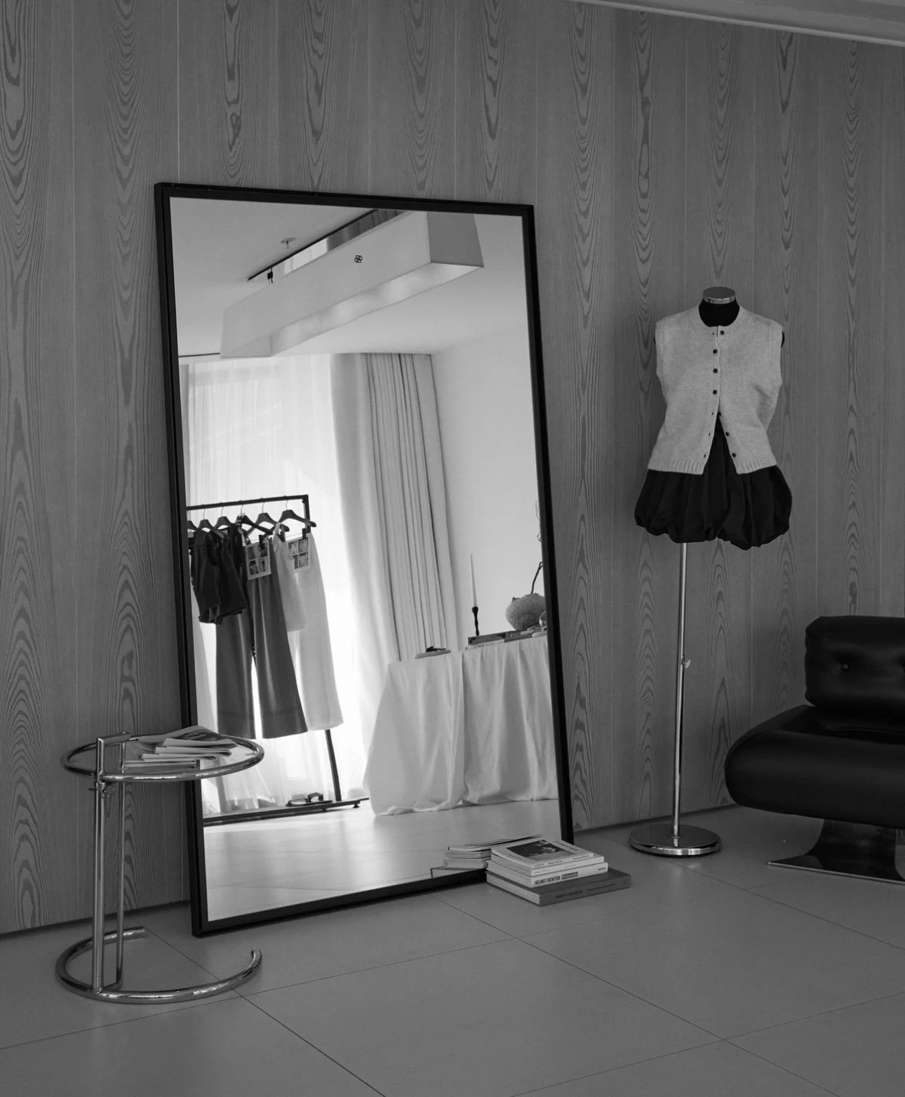 A black and white photo of a fitting room area in a boutique or clothing store. The space features a large standing mirror, a mannequin dressed in a vest and puffy skirt, a small round table with magazines, a clothing rack with garments, and a table 
