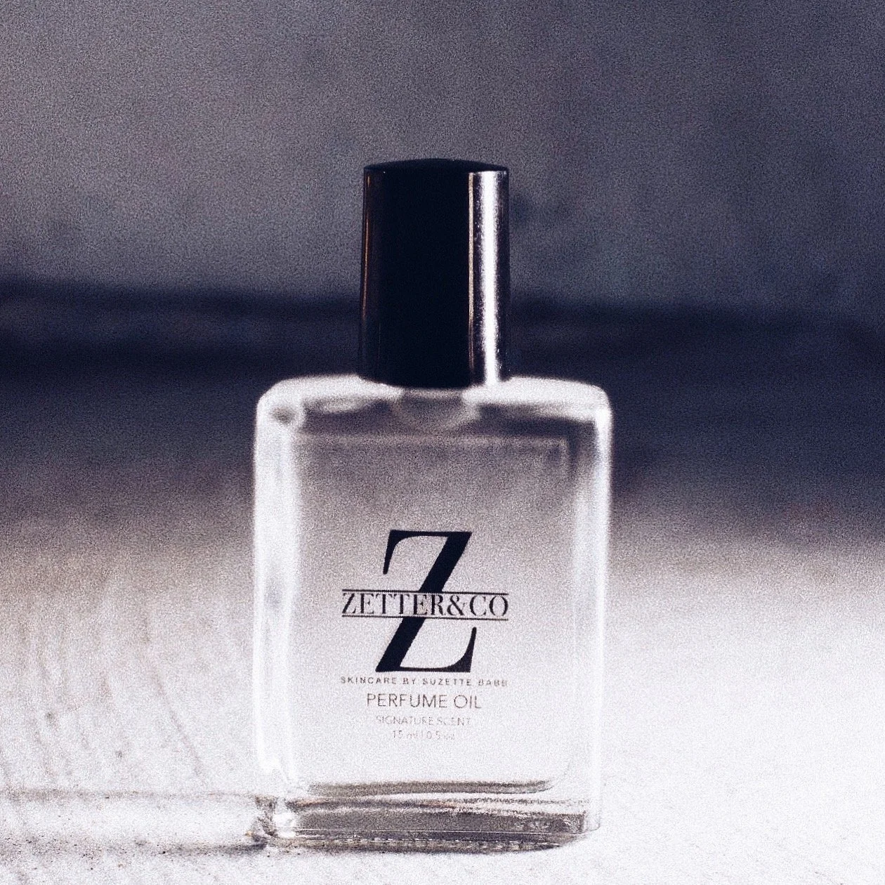 A clear glass perfume oil bottle with a black cap, labeled Zetter & Co by Suzette Dunn, on a light-colored surface with a gradient background.