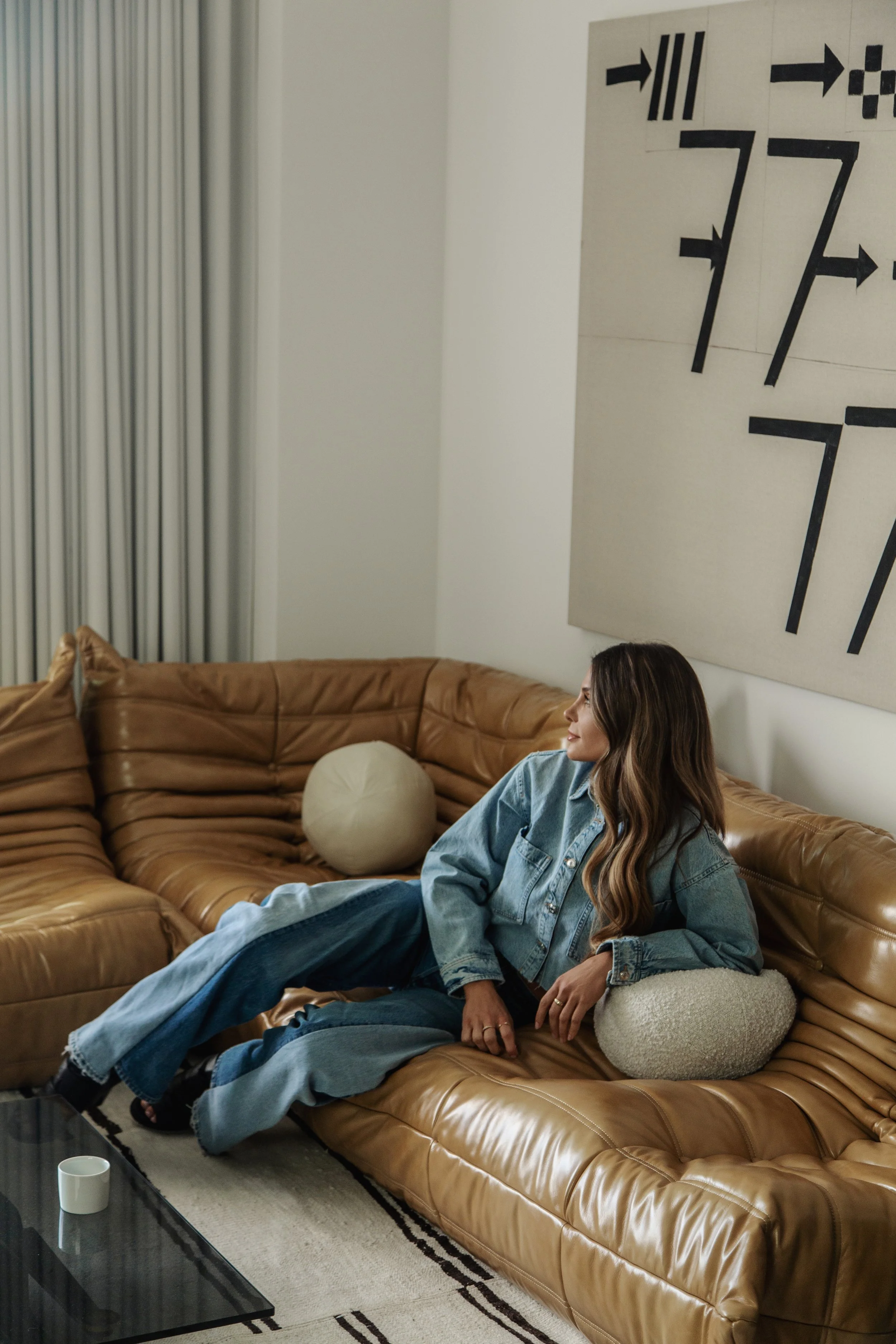 A woman with long, wavy brown hair, wearing a light blue denim jacket and blue jeans, sitting on a tan leather sectional sofa in a living room, smiling and looking to her left. There are beige pillows behind her, a small round beige pillow on her lap