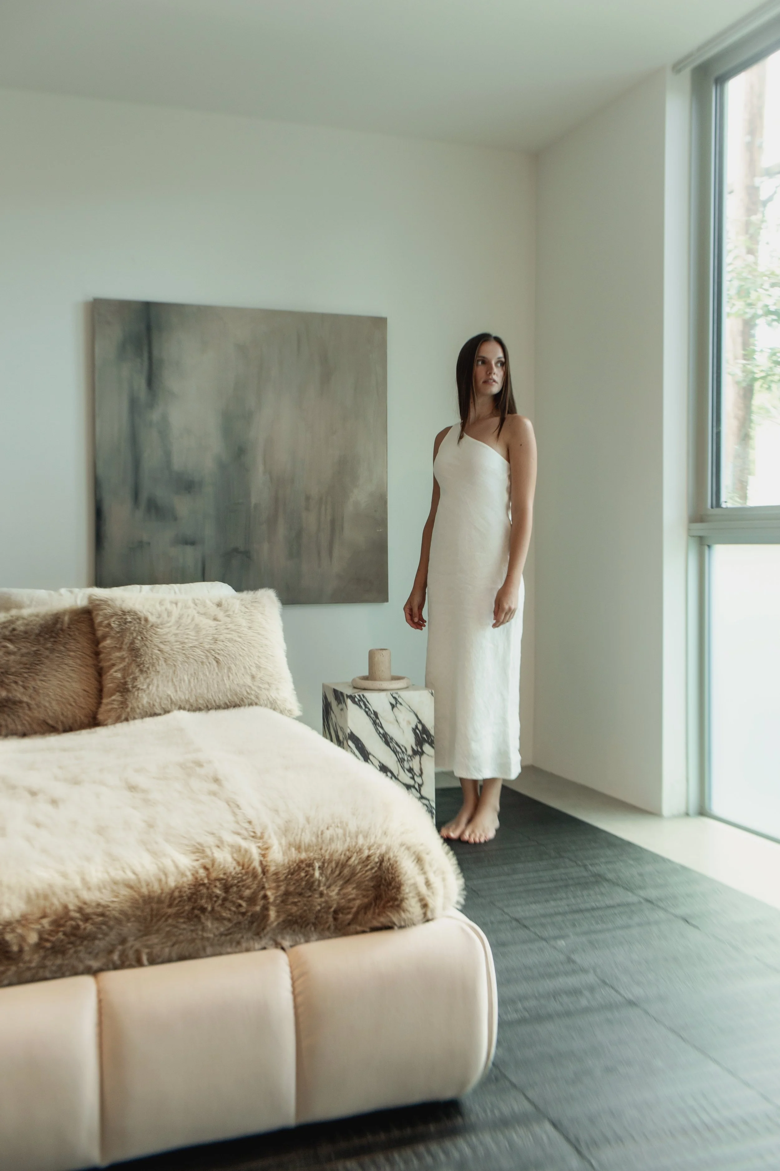 A woman in a white dress standing barefoot near a large window in a modern, minimalist living room with a fur-covered sofa, a marble side table with a candle, and artwork on the wall.