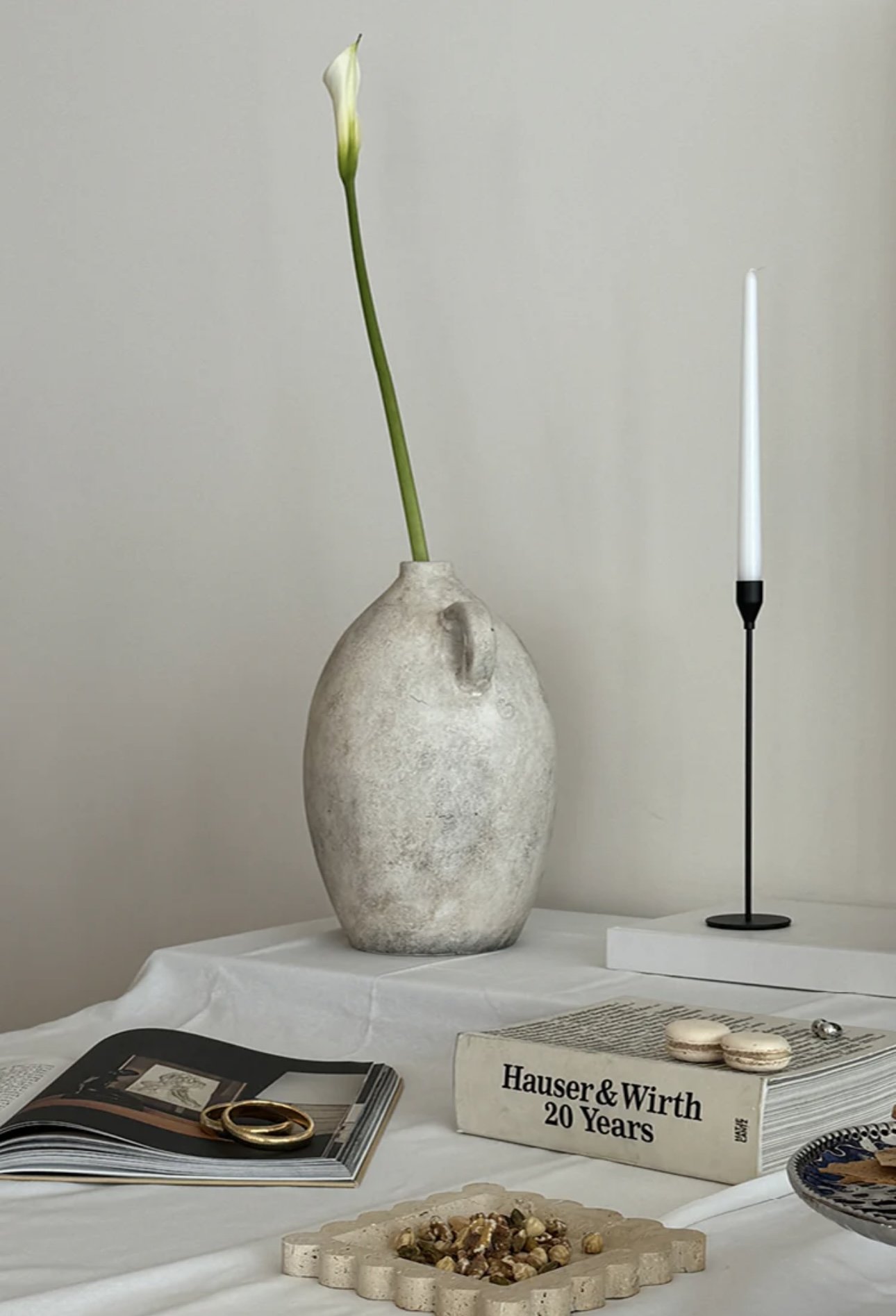White textured vase with a single green and white flower stem, a narrow lamp on a white base, open magazine with gold ring, box labeled "Hauser & Wirth 20 Years," plate with nuts, small round object, and a piece of paper on a white tablecloth.