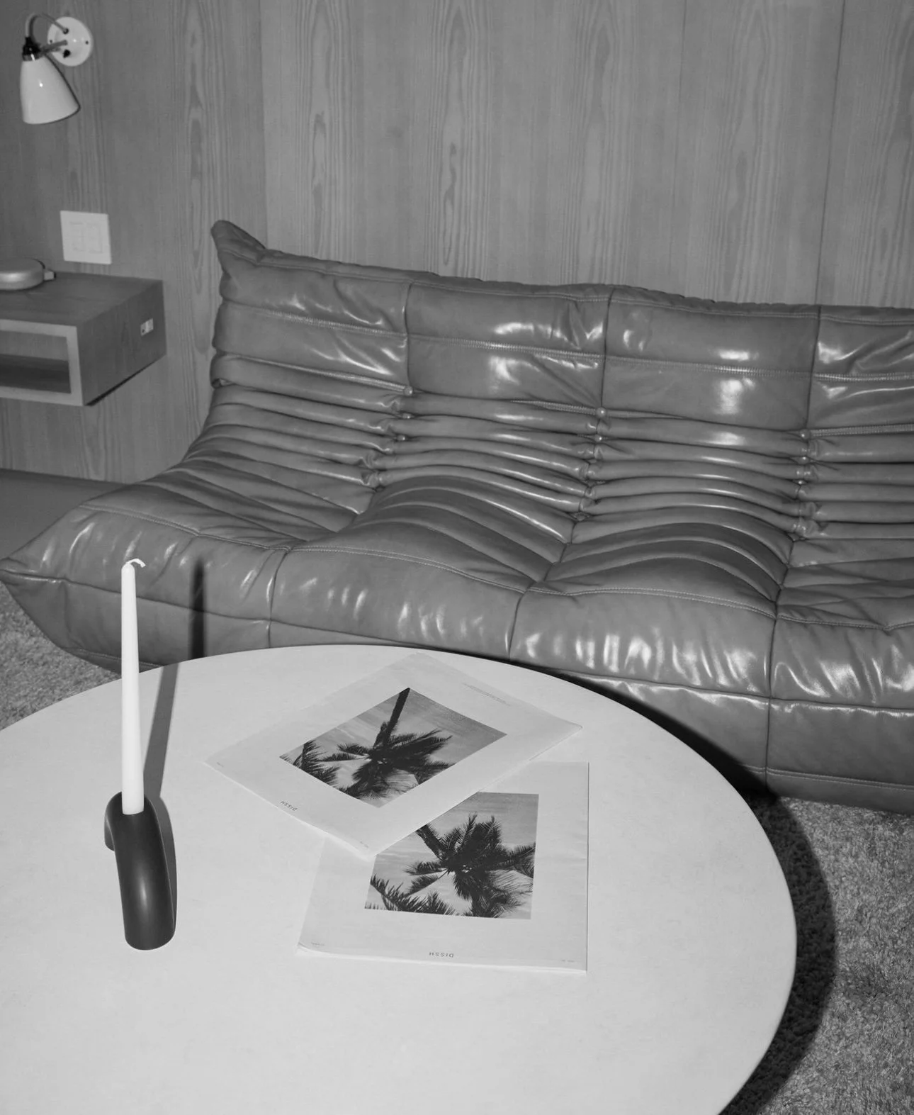 A living room with a curved leather sofa, a round table with a candle, and two palm tree photographs on the table.