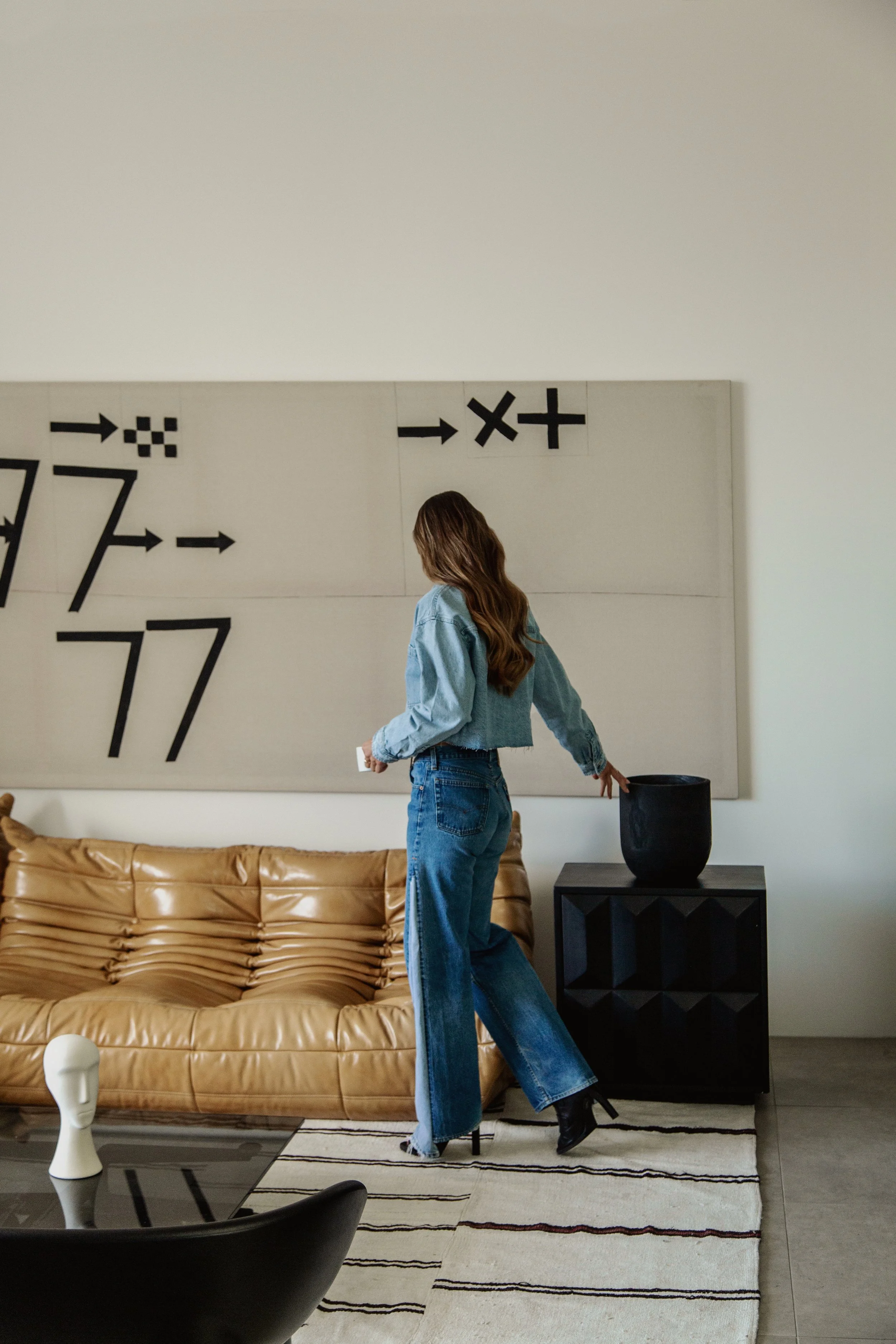 A woman with long hair wearing a blue denim jacket, blue jeans, and black boots is in a modern living room. She is walking past a black side table with a large black vase on it, in front of a tan leather sofa and a black-and-white striped rug. There 