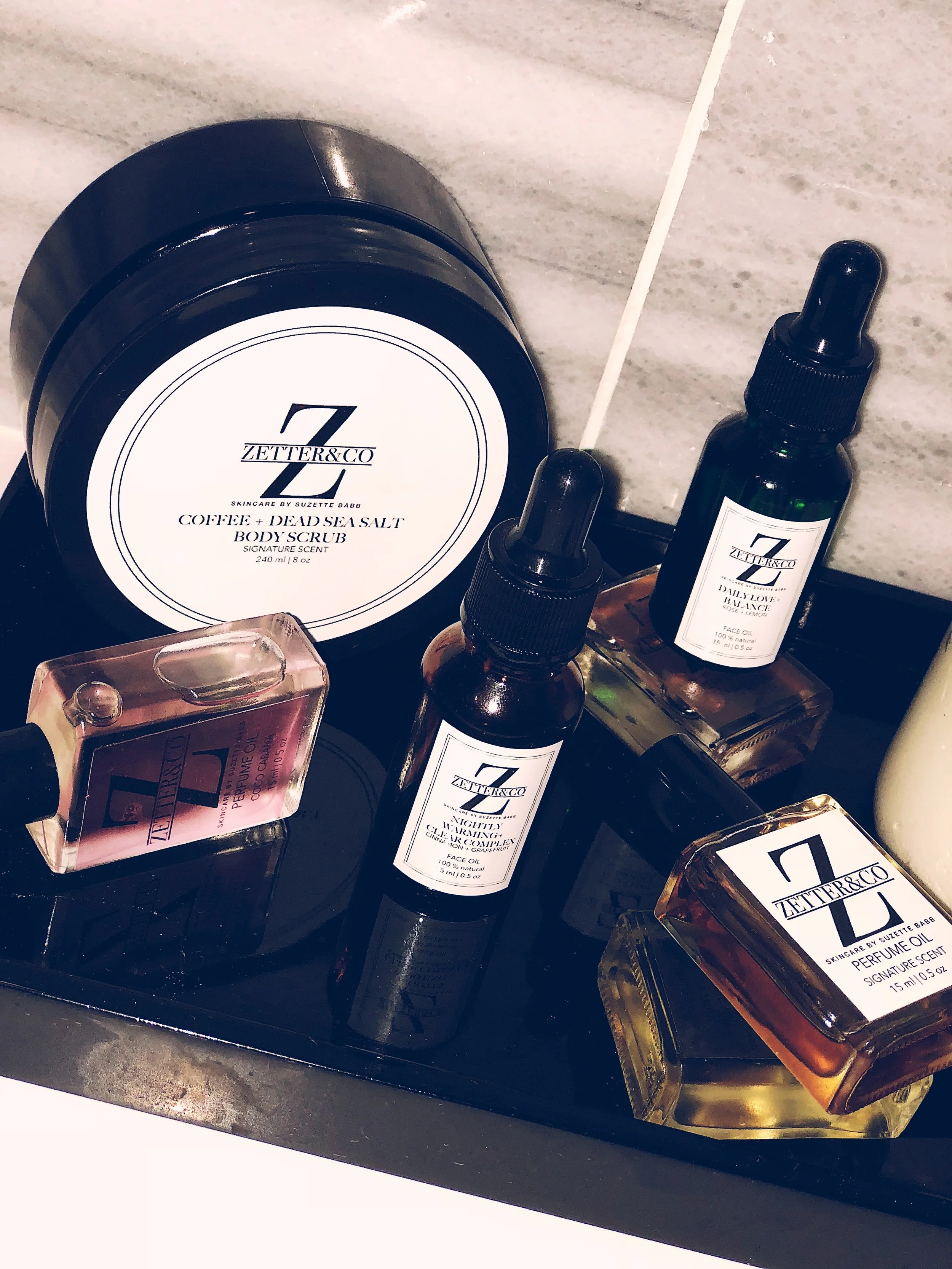 Collection of Zetter&Co skincare products including body scrub and facial oils on a black tray, with a marble surface background.