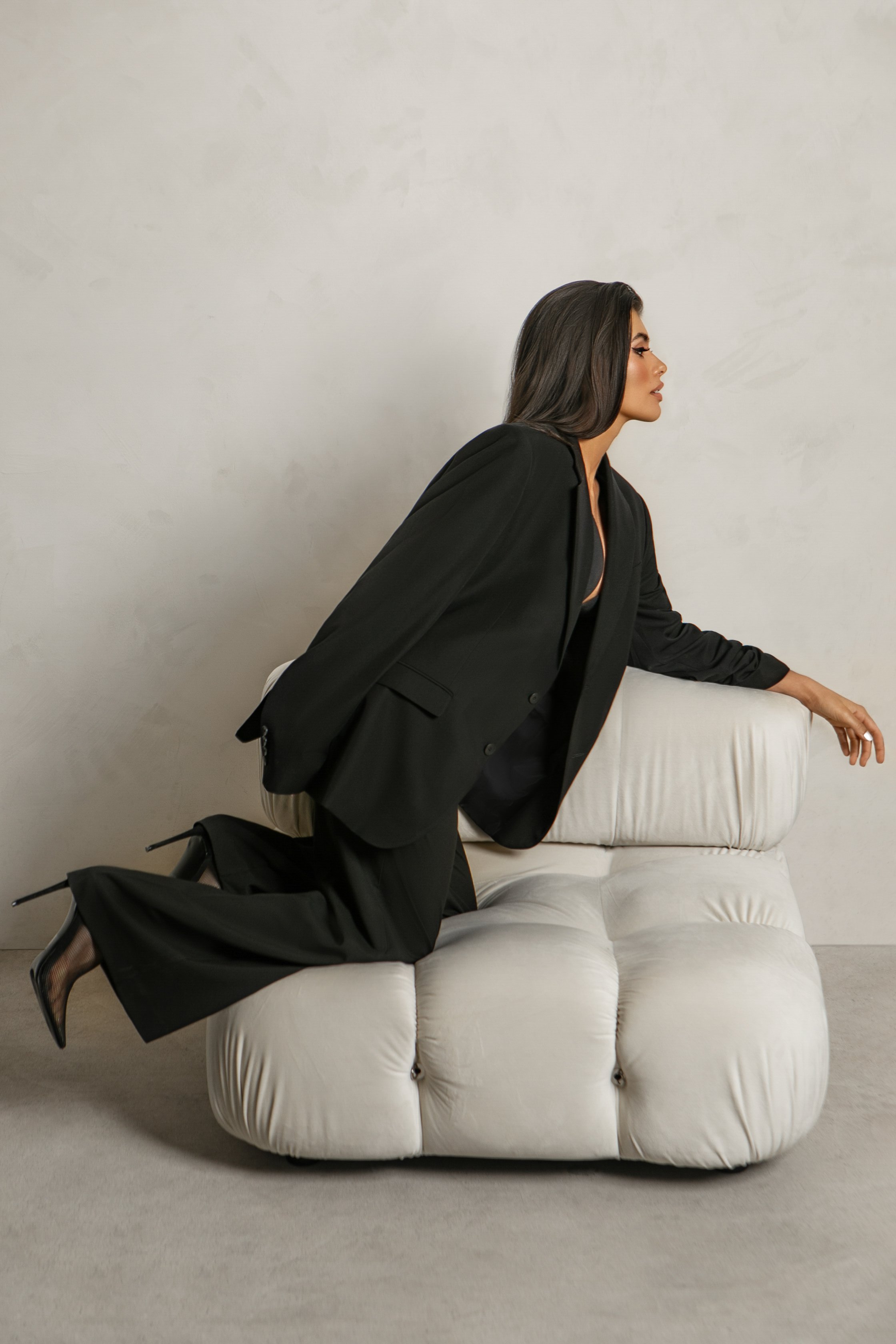 A woman in a black blazer and shoes is kneeling on a cream-colored tufted sofa, facing right with her right arm resting on the sofa's armrest and her left hand on her knee, against a plain light-colored wall.